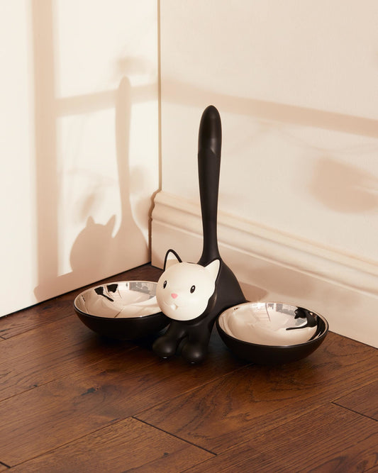 The Tigrito dual cat bowl for food and water features a cartoon black and white cat flanked by two bowls blending black thermoplatic on the outside and stainless steel inner for hygiene. Ideal alternative to boring food bowls for cat owners
